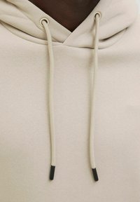 Beige Kapuzenpullover mit Kordelzug, verfügt über eine glatte Textur und schwarze Enden an den Kordeln. Einfaches Design ohne Muster.