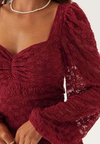 Gros plan sur une personne portant une robe en dentelle bordeaux avec des manches bouffantes et un collier de perles.