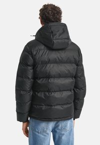 Man i svart puffjacka med huva och blå jeans, sedd bakifrån mot en enfärgad bakgrund.