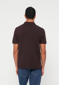 Tmavě hněré polotriko s krátkými rukávy, plochým límcem a rovnou spodní linií, kombinované s modrými džínami. Hladká textura látky.