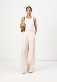 Top de tirantes blanco con canalé combinado con pantalones de pierna ancha en un tono beige claro. Un bolso marrón cuelga a su lado. Tejido suave con una textura sutil.