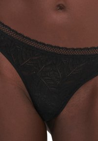 String noir en dentelle avec un motif floral et une taille festonnée. Le tissu est semi-transparent et a une texture lisse.