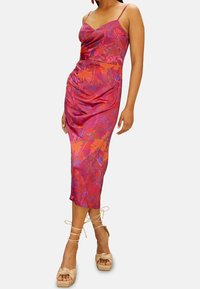 Nainen pukeutuneena vaaleanpunaiseen ja oranssiin kukalliseen hihattomaan midi-mittaiseen mekkoon, jossa on ohuet olkaimet, ja beigeihin nilkkasilmukkasandaaleihin, joissa on korkeat korot.