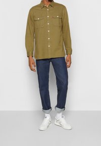 Oliwkowa koszula z guzikami z dwiema kieszeniami na piersiach, zestawiona z ciemnoniebieskimi dżinsami i białymi trampkami, eksponowana z podwiniętymi rękawami.