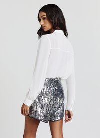 Blusa bianca a maniche lunghe con colletto e cucitura posteriore sottile, abbinata a pantaloncini argento scintillanti con paillettes e vita arricciata.