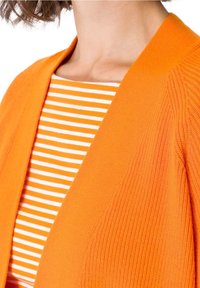 Oranžový pletený cardigan s texturovanou látkou, s otevřeným předním designem, nošený přes bílou a oranžovou pruhovanou košili.