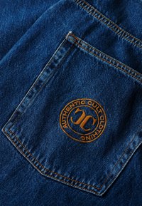 Tela de mezclilla de color azul oscuro, con costuras naranjas y un logo bordado circular en el bolsillo trasero que dice "AUTÉNTICA ROPA CULTURAL."