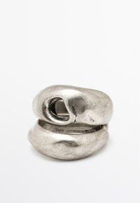 Anello in metallo con forme scultoree irregolari di colore argento. Presenta una superficie texturizzata e un'apertura nella parte superiore dell'anello.