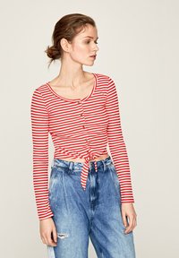 Maglietta a maniche lunghe a righe rosse e bianche con scollatura rotonda, bottoni frontali e orlo annodato. Abbinata a jeans in denim blu a vita alta.