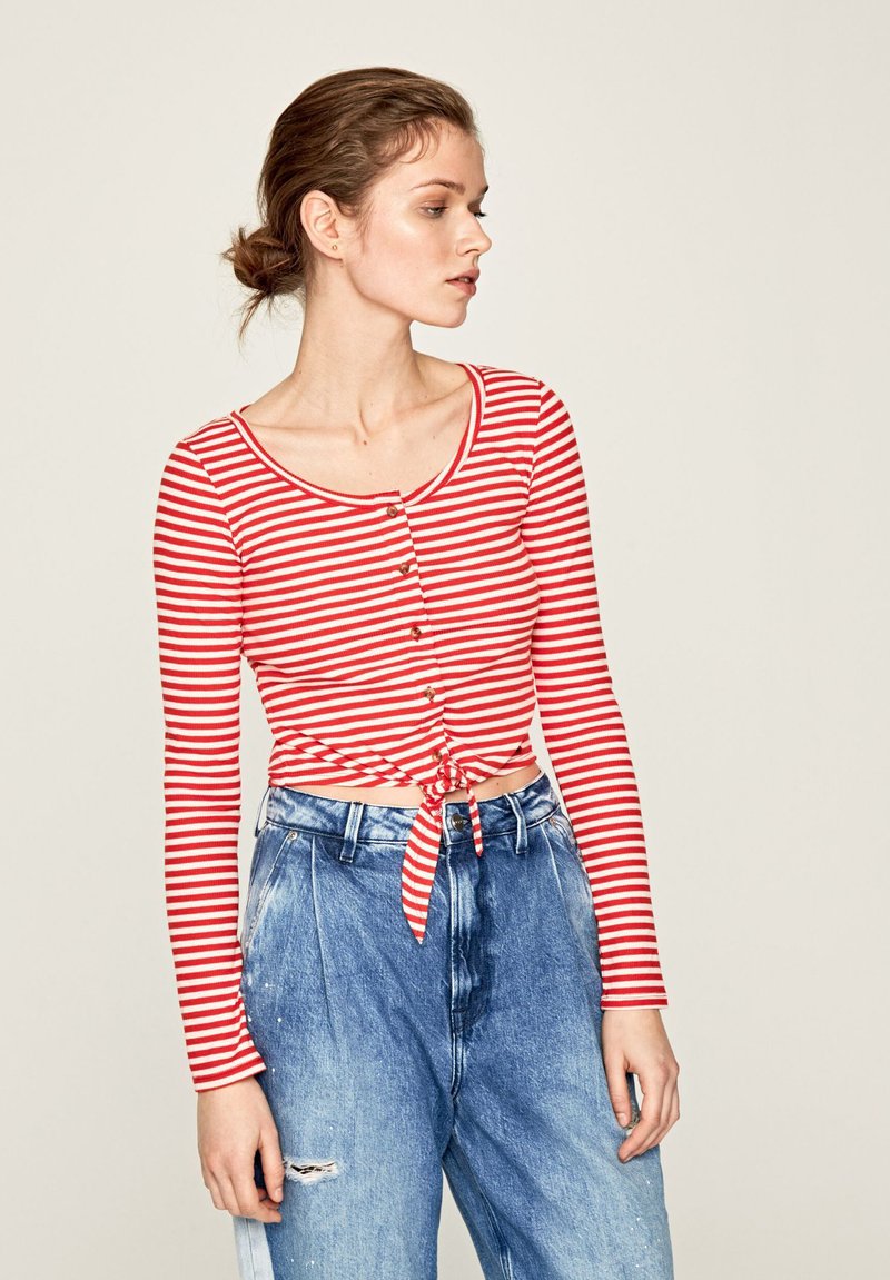 Maglietta a maniche lunghe a righe rosse e bianche con scollatura rotonda, bottoni frontali e orlo annodato. Abbinata a jeans in denim blu a vita alta.