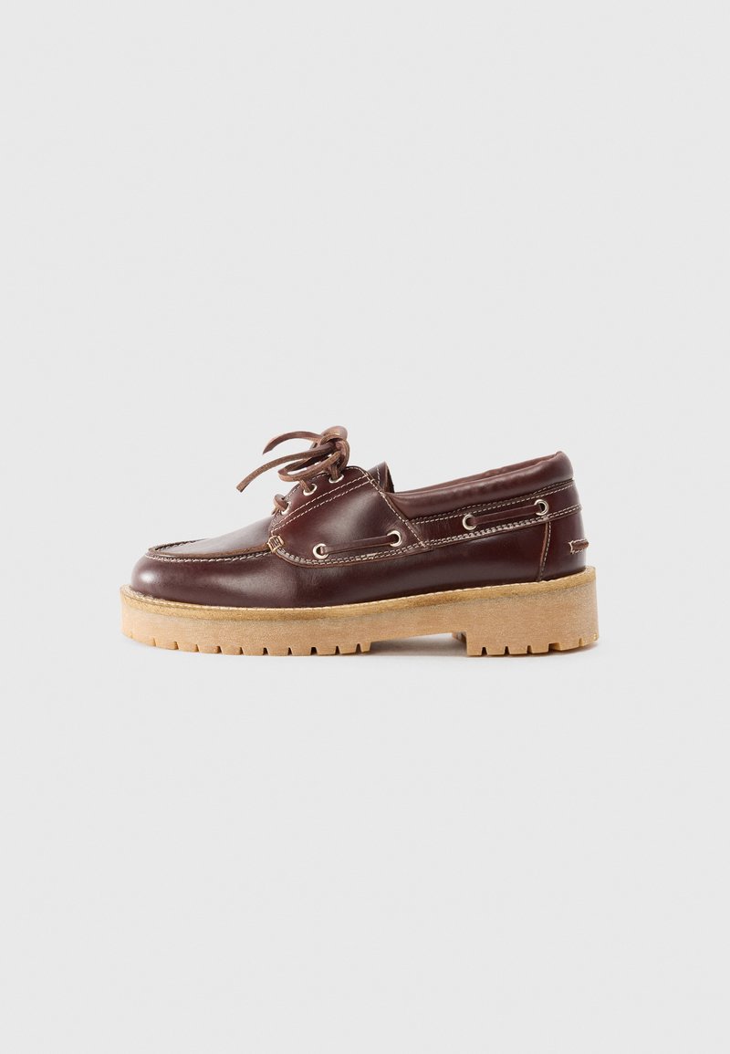 Zapato de barco de piel marrón con puntera redonda, parte delantera con cordones y ojetes de metal, que presenta una suela gruesa de color marrón claro y costuras en contraste.