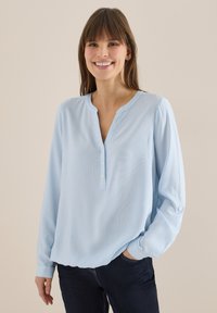 Femme souriante aux cheveux bruns portant un chemisier à manches longues rayé bleu clair et blanc, ainsi qu'un jean foncé, debout devant un fond beige uni.