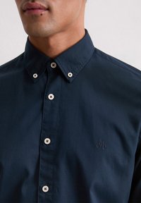 Marineblauw overhemd met knoopsluiting en een puntige kraag, voorzien van witte knopen en een gegraveerd logo. Gladde textuur, op maat gesneden pasvorm en geen zichtbare patronen.