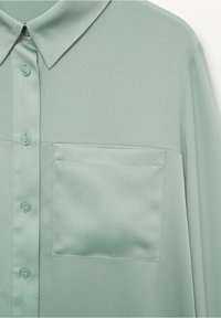 Devant de chemise en satin vert clair avec col, patte de boutonnage et poche poitrine, montrant une texture de tissu lisse et légèrement réfléchissante.