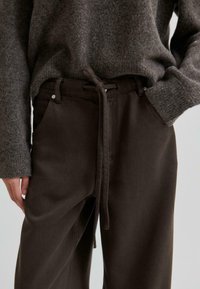 Pantaloni in cotone marrone con vita regolabile e tasche laterali, abbinati a un maglione di maglia grigia. La texture sembra morbida e accogliente.