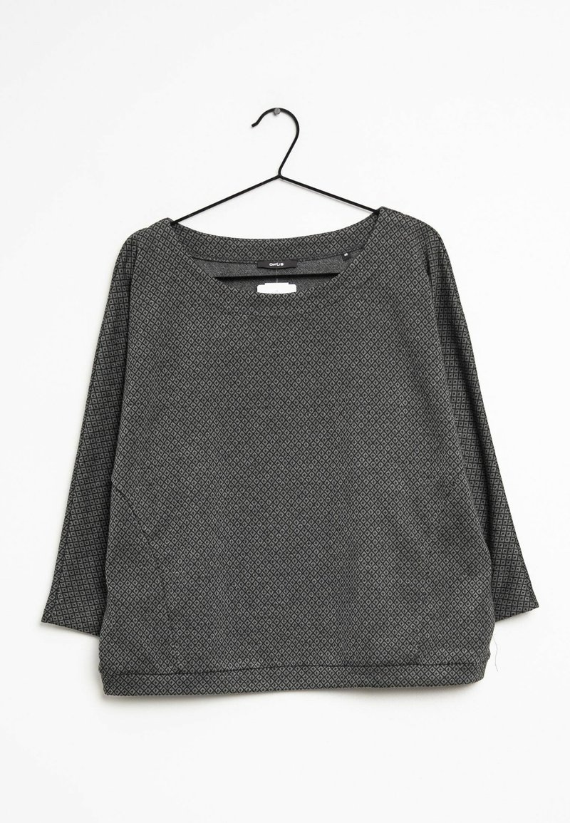 Camisa de manga larga de patrones en negro y gris con un escote amplio y un corte holgado, colgando de una percha de metal negro sobre un fondo blanco.