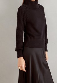 Schwarzer Rollkragenpullover mit strukturierten, gerippten Ärmeln und einer taillierten Passform. Kombiniert mit einem seidenen schwarzen Rock. Elegantes Strickdesign mit einer strukturierten Silhouette.