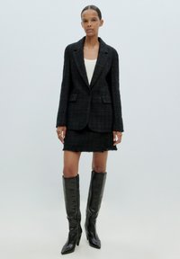 Schwarzer strukturierter Blazer mit Revers, einem Ein-Knopf-Verschluss, kombiniert mit einem passenden mini-rock mit Fransen und knielangen schwarzen Lederstiefeln.