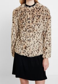 Tekoturkista valmistettu takki vaaleanruskeana, jossa on leopardikuosiset yksityiskohdat. Takissa on pitkät hihat ja pyöreä kaula-aukko. Pehmeä rakenne ja rento istuvuus.