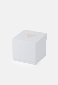 Balta dėžutė su tekstūruotu paviršiumi, ant kurios viršaus uždėtas aukso trikampio logotipas, kvadrato formos ir nuimama dangteliu. Paprastas ir minimalistinis dizainas.