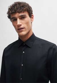 Camicetta nera con colletto classico, dotata di bottoni argentati e tessuto liscio. Il design è sartoriale con una silhouette aderente.