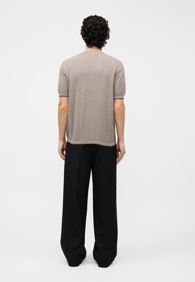 Άνδρας με σγουρά μαύρα μαλλιά στέκεται με την πλάτη προς τα εμπρός, φοράει μπεζ κοντομάνικο πουκάμισο με υφή και φαρδιά μαύρα παντελόνια σε απλό φόντο.