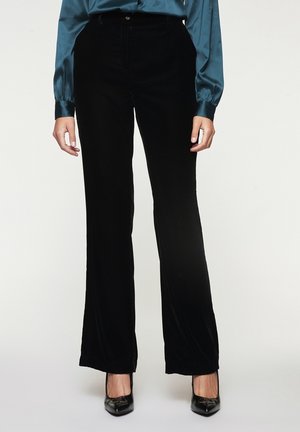 Persoon die zwarte broek met wijde pijpen draagt, een teal satijnen blouse met lange mouwen en zwarte puntige hakken, staand tegen een effen achtergrond.