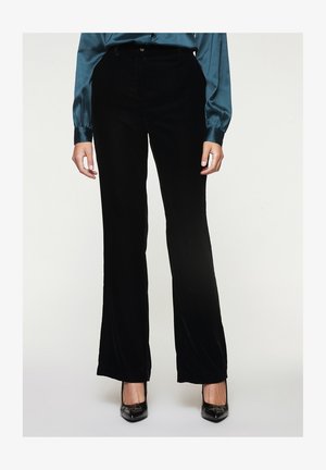 Person iført sorte bukser med brede ben, teal satin langærmet bluse og sorte spidse hæle, stående mod en ensfarvet baggrund.