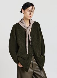 Cardigan oversize verde scuro con scollo a V e tre bottoni, abbinato a una sciarpa beige chiaro annodata attorno al collo. Texture liscia, vestibilità casual.