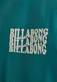 Teal cotton t-shirt featuring repeated white "BILLABONG" text design in a wavy font. The fabric has a soft texture.