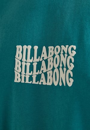 Teal cotton t-shirt featuring repeated white "BILLABONG" text design in a wavy font. The fabric has a soft texture.