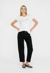 T-shirt blanc avec l'inscription "Liu Jo" en texte pailleté argenté, associé à un pantalon noir ample avec des coutures décoratives et un bouton à l'avant.