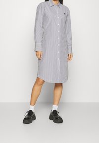 Donna che indossa un vestito a righe con maniche lunghe e bottoni, lungo fino al polpaccio, con calzini bianchi e scarpe con plateaux nere, in piedi contro uno sfondo neutro.