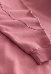 Close-up van een roze sweatshirtmouw met geribde manchetten en gebogen naadstiksels op zachte stof.