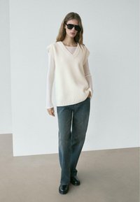 Kremfarvet strikket ærmeløs sweater med V-hals, båret over en langærmet top, kombineret med mørke denimjeans og sorte ankelstøvler.