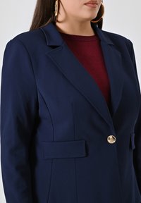 Marineblauwe blazer met een enkele gouden knoop, twee voorzakken en scherpe revers. Draagt over een bordeaux rode gebreide top. Gladde stoftextuur.