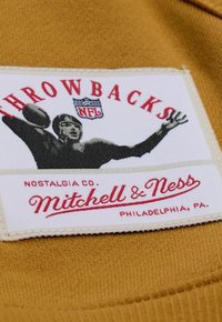 L'etichetta dell'abbigliamento vintage NFL presenta un'etichetta in tessuto bianco con testo rosso, un'immagine di un giocatore che lancia un pallone da football e un materiale di fondo color beige.