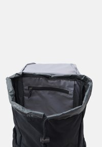 Sac à dos en tissu noir avec un intérieur gris. Doté d'une fermeture à cordon et d'une poche zippée externe sur un côté.