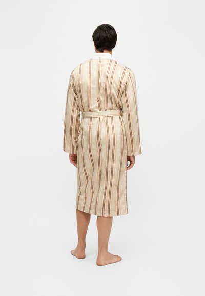 Homme debout pieds nus portant un peignoir beige avec des rayures verticales marron, vu de dos sur un fond blanc uni.