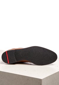 Suola di scarpa in pelle marrone con gomma nera testurizzata, caratterizzata da una striscia rossa di accentuazione sul tallone. Costruzione robusta progettata per la durata.