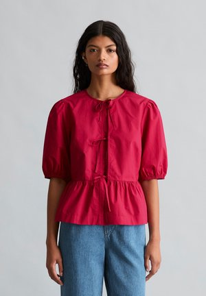 Femme aux longs cheveux foncés portant un chemisier rouge à manches bouffantes avec des fermetures à nouer et un jean bleu taille haute, debout devant un fond gris uni.