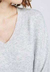 Pull à col V gris clair en tricot doux et texturé. Il présente un col côtelé et une légère ampleur au niveau des manches.