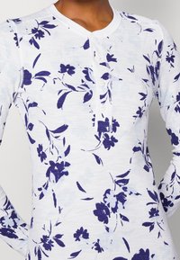 Haut à manches longues en blanc, avec un imprimé floral marine. Tissu texturé avec un col Henley à boutons et des poignets côtelés.