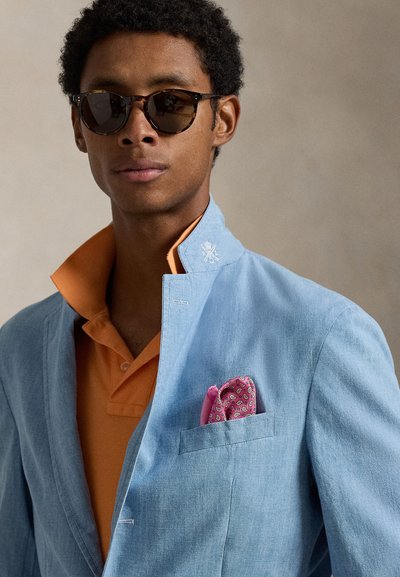 Ανοιχτό μπλε λινό σακάκι με κλειστό γιακά, σε συνδυασμό με πορτοκαλί πόλο πουκάμισο. Διαθέτει ένα ροζ μαντίλι με σχέδια στην τσέπη στο στήθος.