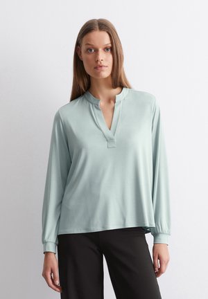 Blusa verde chiaro a maniche lunghe con colletto alla mandarina e pattina a bottoni. Texture liscia, vestibilità comoda e polsini arricciati.