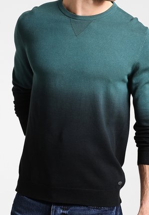 Mann trägt einen Langarm-Pullover mit Rundhalsausschnitt in einem Farbverlauf von Türkis zu Schwarz, kombiniert mit blauen Jeans.