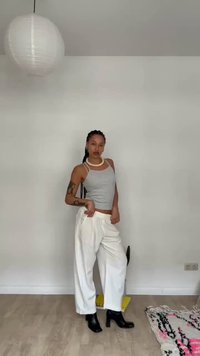 Mujer con cabello trenzado que lleva una camiseta sin mangas gris, pantalones blancos de pierna ancha, botas negras con tacón, de pie en una habitación minimalista con una lámpara colgante.