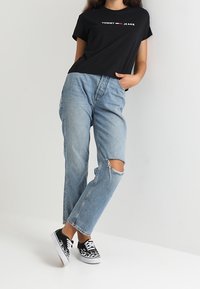 Camiseta de algodón negra con el logo "TOMMY JEANS". Jeans de cintura alta en azul claro con una rodilla desgastada. Zapatillas negras con cordones blancos y patrón.