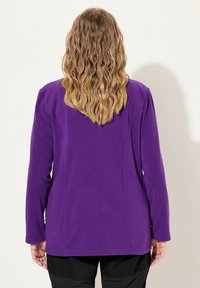 Donna con capelli biondi ondulati, che indossa una giacca viola e pantaloni neri, in piedi di fronte a una parete di colore chiaro, con le mani rilassate ai lati.