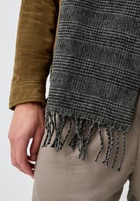 Écharpe en laine à carreaux gris avec bords frangés, drapée sur une veste en daim marron. L'écharpe présente un design texturé et des fils de couleurs contrastées.